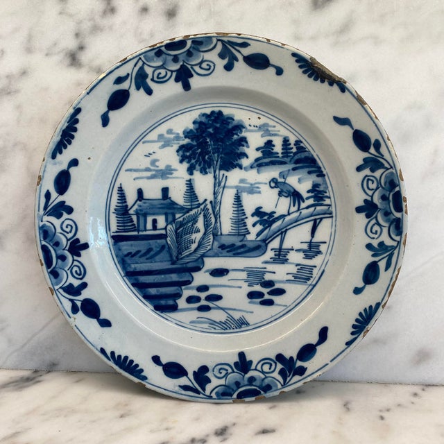 A beautiful antique Dutch Delft blue and white plate, hand-painted with a picturesque riverside landscape featuring a...