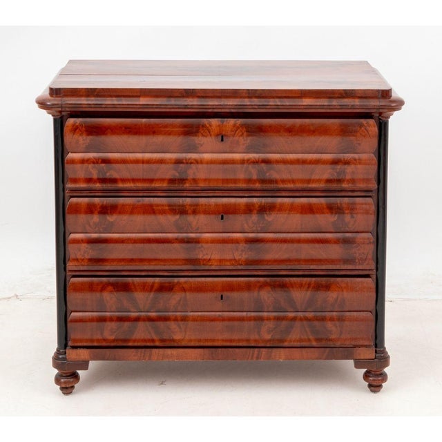 Biedermeier Flame Mahogany Commode, the ogee facade with three drawers flanked by ebonized columns. 36" H x 40.5" W x...