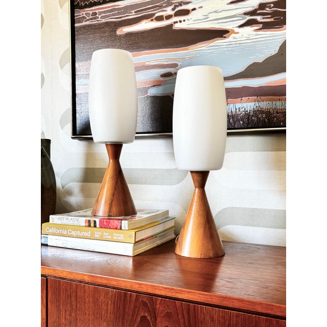 A pair of matching mid century table lamps. Each features a wood base and frosted glass shade. Bases are a medium toned...