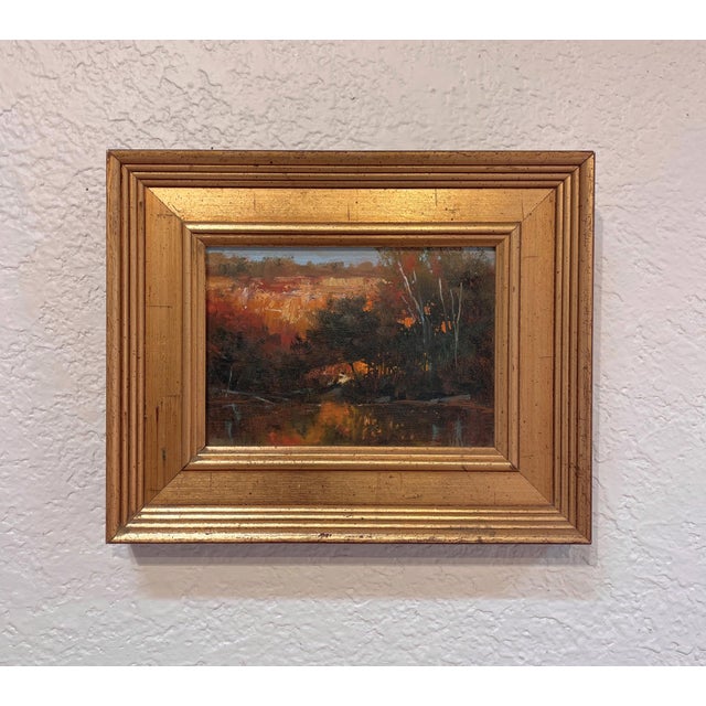 This oil on panel artwork captures a serene river scene bathed in warm autumn hues, reflecting the rich colors of the...