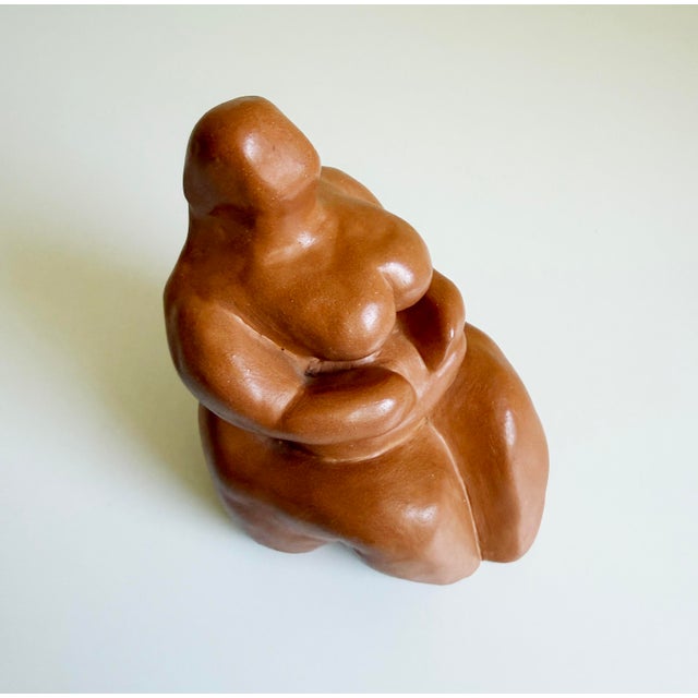 An unglazed red-brown clay sculpture depicting a highly stylized, voluminous female form in a seated position. The...