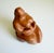 An unglazed red-brown clay sculpture depicting a highly stylized, voluminous female form in a seated position. The...