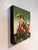 This artwork depicts a vibrant bird perched atop a whimsical arrangement of assorted objects, including colorful flags and...