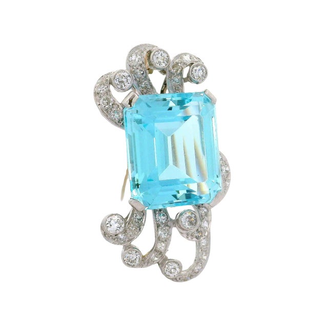A stunning brooch showcasing a mesmerizing emerald-cut aquamarine, weighing 14.00 carats, in a vivid blue hue. The...