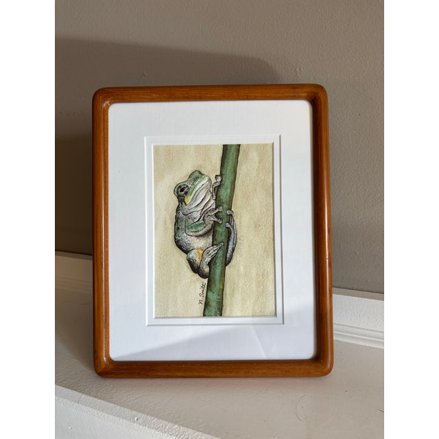 A small frog clings quietly to a vertical reed, captured in exquisite detail and earthy watercolor tones. The textured...