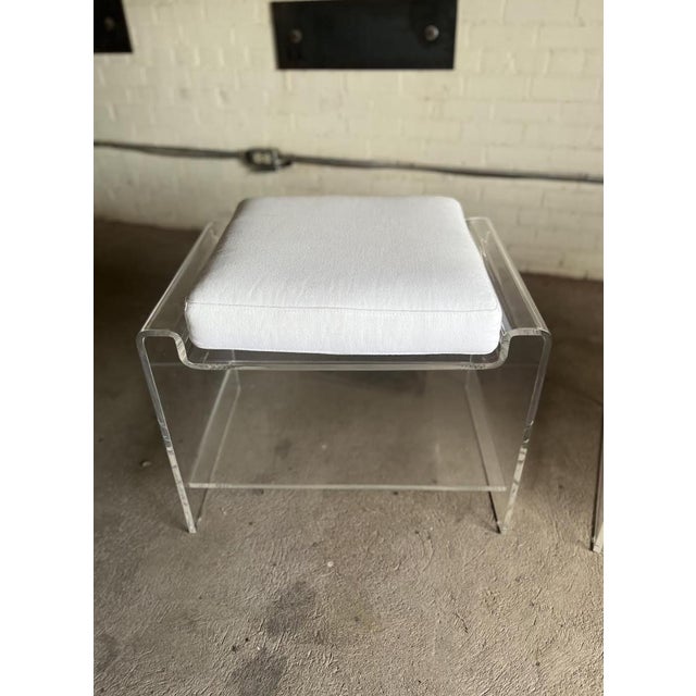 A stylish pair of vintage Lucite waterfall ottomans featuring thick clear acrylic frames and crisp white upholstered...