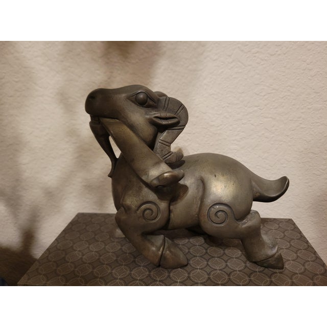 A striking solid metal ram sculpture featuring stylized curves, engraved detailing, and a bold, compact form. The raised...