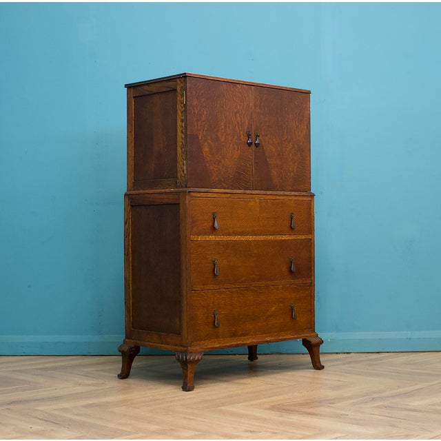 A 1930s Art Deco oak tallboy with a cupboard top section over three drawers, raised on carved shaped feet. The upper...