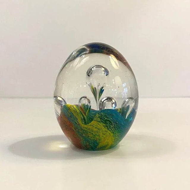 Oval shaped, artisan-made, hand-blown, Italian Murano art glass paperweight features a bed of colorful teal, blue, yellow,...
