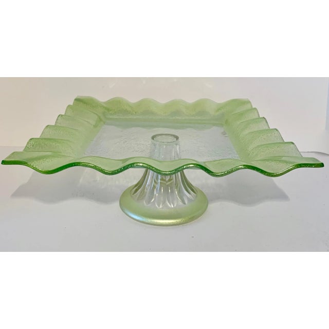 A square glass footed cake or pastry display plate with glass 'Ruffle' around the perimeter. The footed plate is made of...