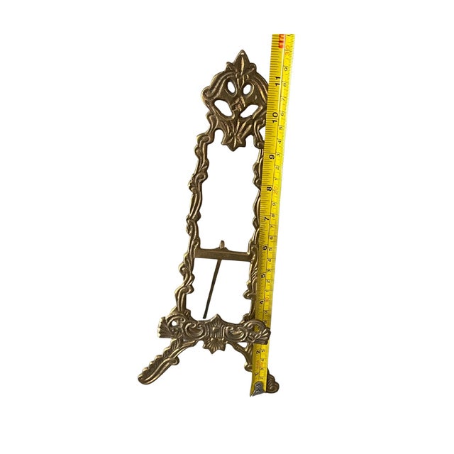 It is crafted from brass and features intricate, ornate floral and scroll detailing. The easel has a folding leg on the...