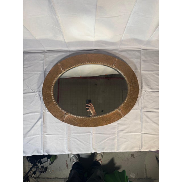 An oval wall mirror in the British Arts and Crafts manner: a hammered metal frame in a warm copper-to-bronze tone with...