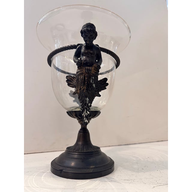 This decorative piece features a clear glass vessel mounted on a bronze pedestal base, adorned with a detailed cherub...