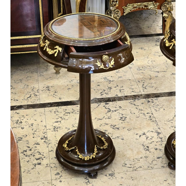 A refined French Neoclassical side table crafted with a rich dark wood finish, a glass-inset tabletop, and delicate gold...