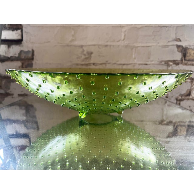 Avocado green hobnail glass bowl with molded feet and a wide, shallow shape. The textured surface and garden-inspired tone...