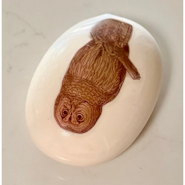 This vintage ceramic paperweight, shaped like an oval egg. features a detailed brown ink illustration of a barred owl...