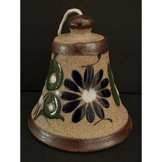 Beautiful handcrafted & hand painted Mexican Folk Art bell adorned with a blue flower and greenery against a specked brown...