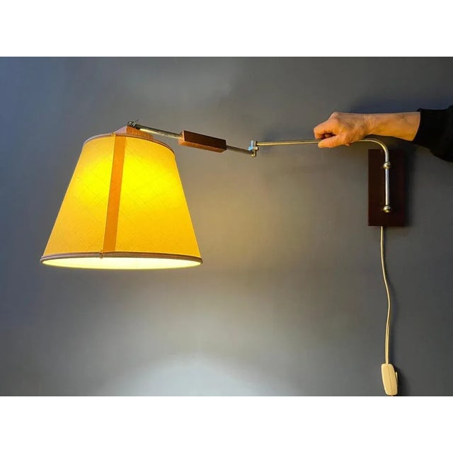 Classy mid-century wall lamp with textile shade and wooden elements. The lamp has a beige textile shade and the arm...