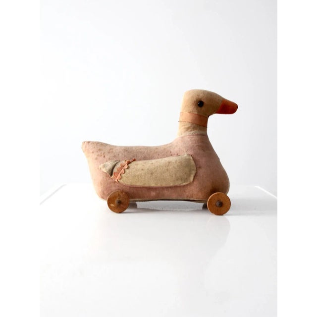 An early 20th century toy duck on wheels. The pink and beige fabric duck features bead eyes, a ribbon with bell around the...