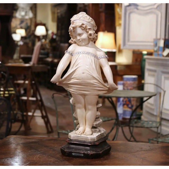 Charming and expressive, this antique plaster sculpture depicts a young girl standing in a playful pose while gently...