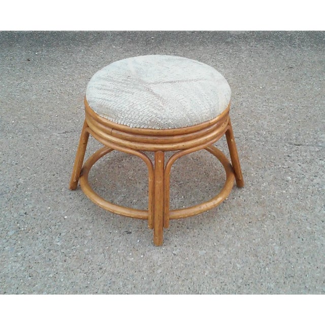 Rattan pouf stool with a medium finish and a cushioned seat upholstered in neutral colored upholstery. This vintage rattan...