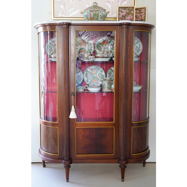 An exquisite Louis XVI style mahogany vitrine featuring a white marble top, fluted corner pilasters, and gilt-bronze trim....