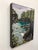This artwork depicts a serene coastal landscape, where vibrant turquoise waters gently lap against rocky shores, framed by...