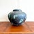 This ceramic vessel features a weathered, volcanic surface with mottled textures of charcoal, ash gray, and oxidized...