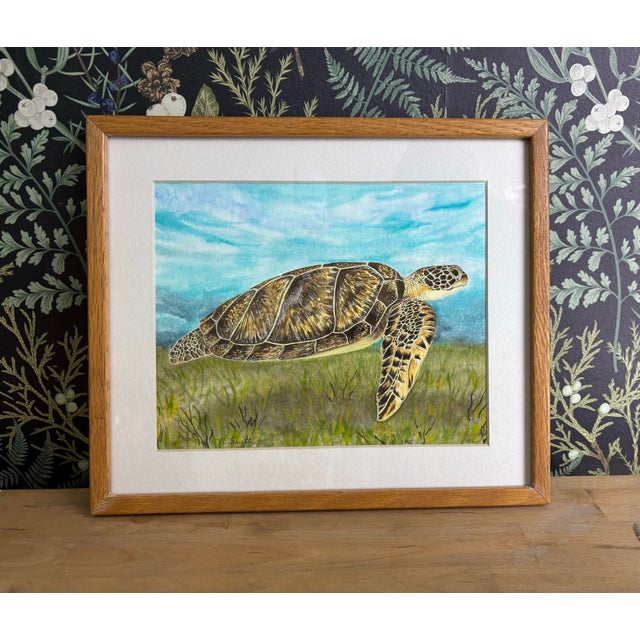 This original watercolor painting features a green sea turtle gliding peacefully over a shallow seagrass bed, rendered in...