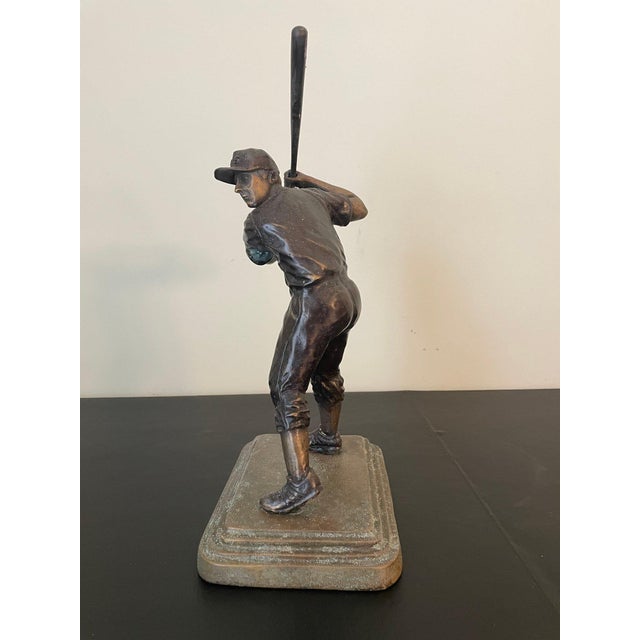 A bronze figural sculpture of a baseball player captured mid-swing, bat raised overhead in batting stance with a period...
