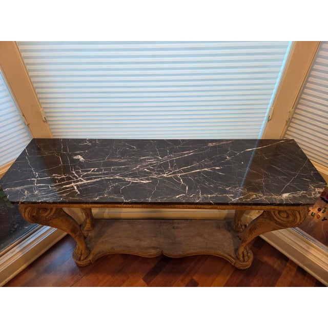 A commanding Neoclassical-style carved wood console table featuring an authentic inset black marble top with striking...