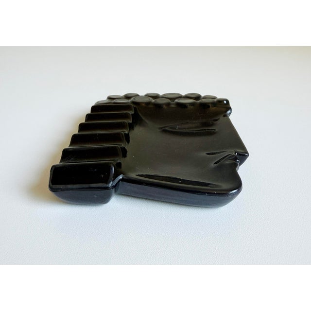 This Art Deco-era ashtray features a stylized male profile and is made of deep black glass with a glossy finish. While...