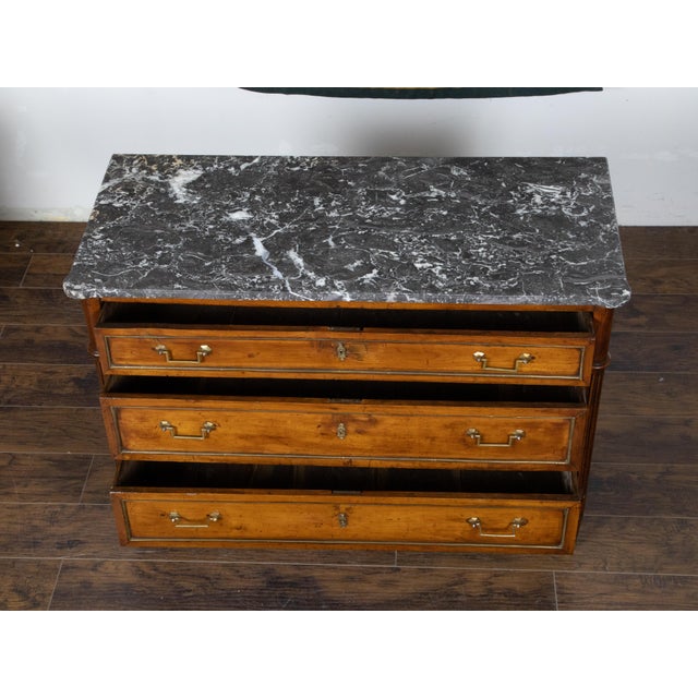 A French Directoire style walnut commode from the 19th century, with three drawers, grey marble top, brass accents and...