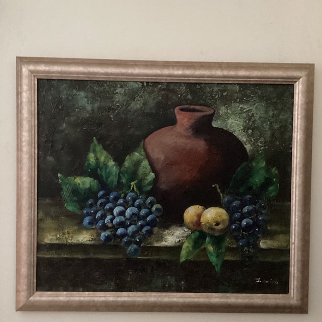 This vintage still life oil painting features a large depiction of fruits and a clay pot, signed by the artist. The...