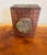 A beautifully aged rectangular tin box featuring a patterned red lid with a raised floral medallion centerpiece. The...