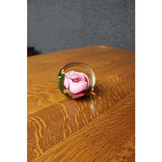 Handcrafted art glass paperweight featuring a sculpted pink rose encased in clear crystal with green accents radiating...
