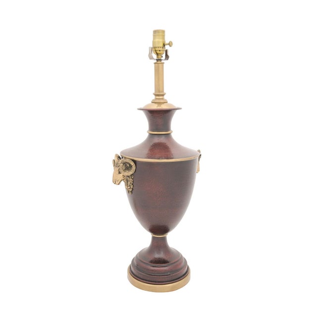 A neoclassical urn-form table lamp featuring ram’s head mounts, rich oxblood textured enamel, and brass accents. Its...