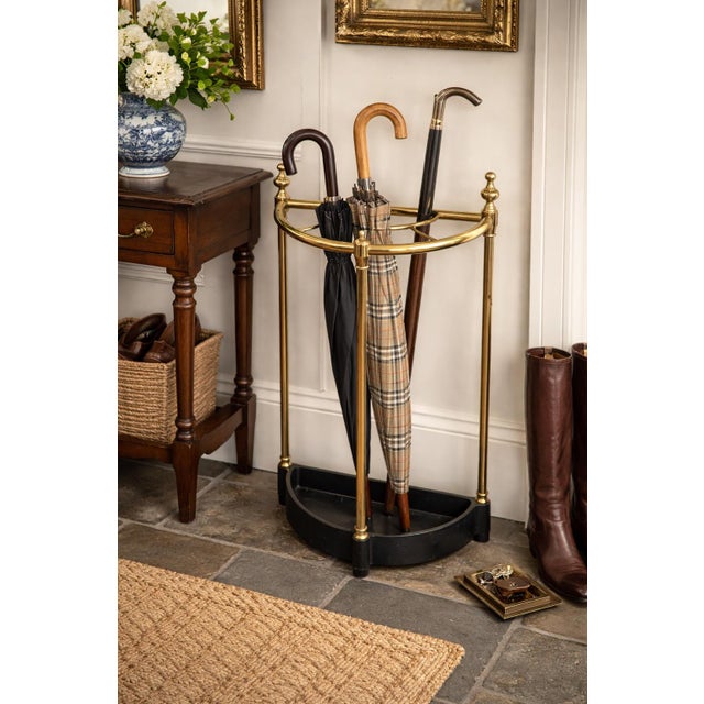 A refined vintage brass umbrella and cane stand featuring a gracefully curved gallery rail with decorative finials and a...