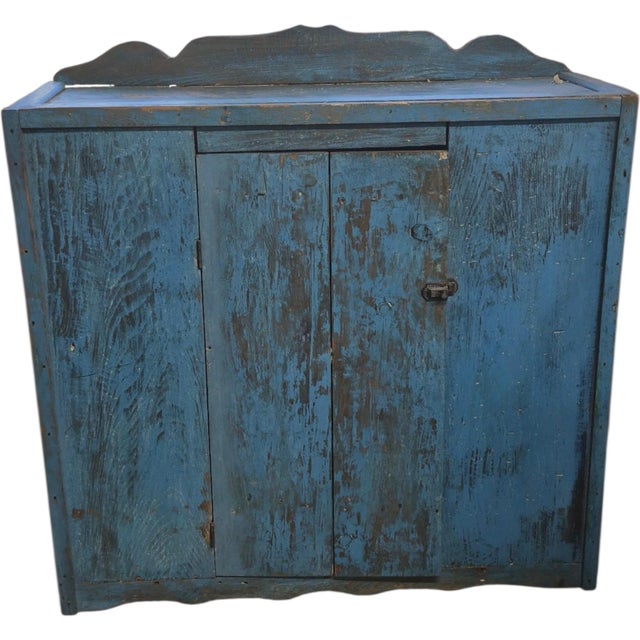A primitive country pine cupboard with a shaped crest, scalloped lower apron, and wide-board door secured by an iron...