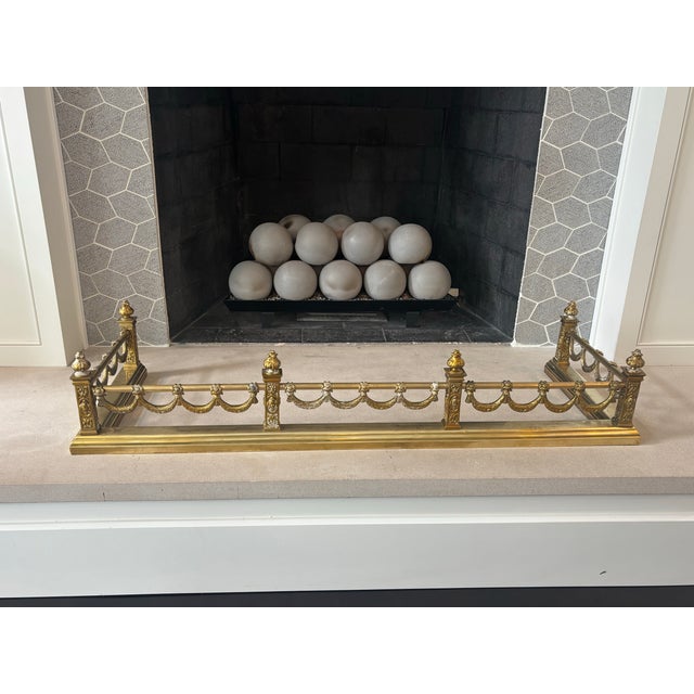 An elegant antique-style brass fireplace fender featuring an ornate neoclassical design with decorative finials, pierced...