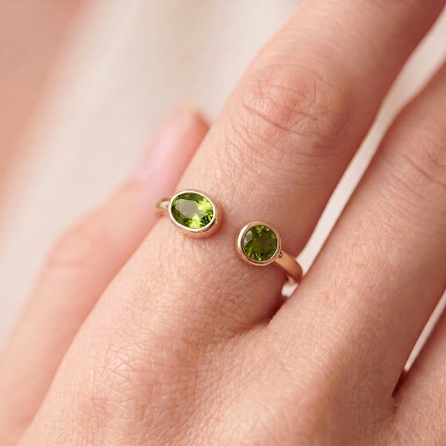 A bold, contemporary open-cuff ring featuring two vibrant green peridots — an oval-cut stone paired with a round-cut stone...