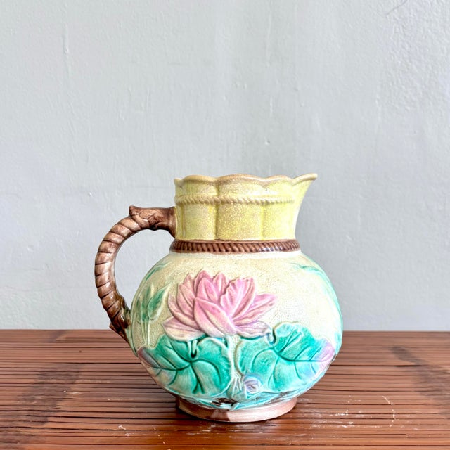This charming ceramic pitcher features a rounded form adorned with raised lotus blossoms and lily pads in soft pastel...