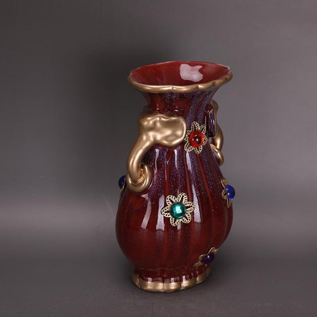 Ornate sculptural ceramic vase with rich oxblood red fluted glaze and subtle purple speckling. It features polished gilt...