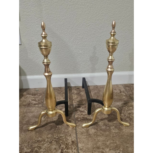 A handsome pair of traditional solid brass andirons featuring elegant turned shafts and timeless urn-form finials. These...