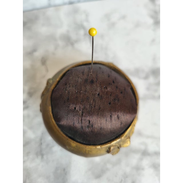 Circa 1900s brassl pin cushion in the shape of a footed ottoman. Minor wear to satin cushion and feet are uneven.