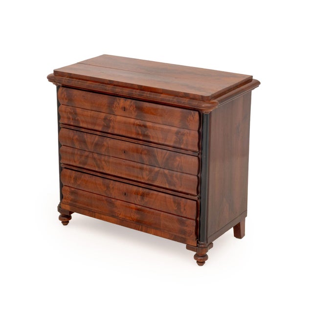 Biedermeier Flame Mahogany Commode, the ogee facade with three drawers flanked by ebonized columns. 36" H x 40.5" W x...