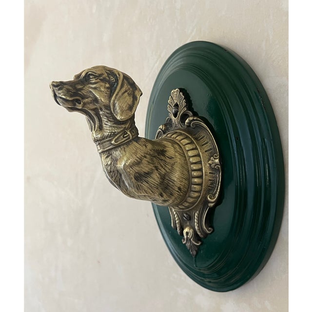 Cast brass dog shaped leash hook on a dark green wooden backplate.