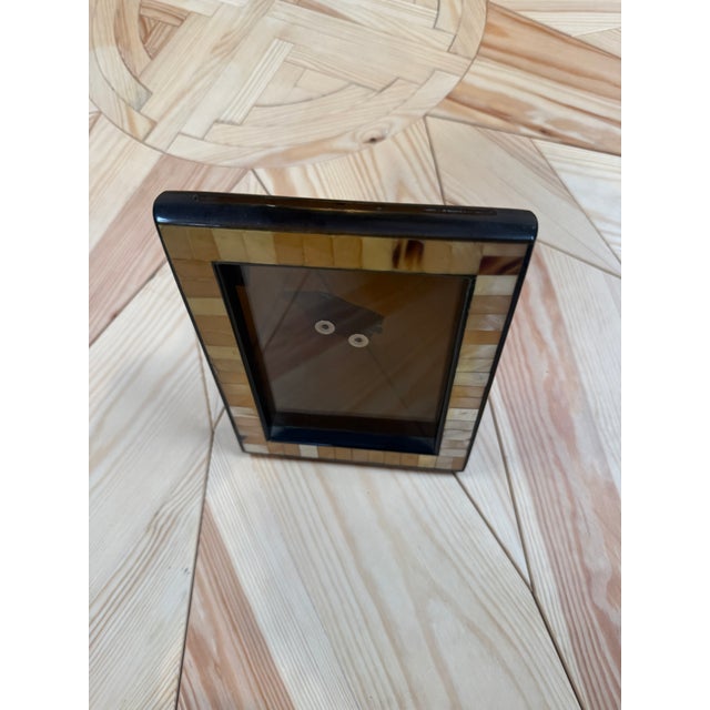 A tabletop picture frame with a border of rectangular tiles in warm cream, honey, and brown tones. Slim dark trim outlines...