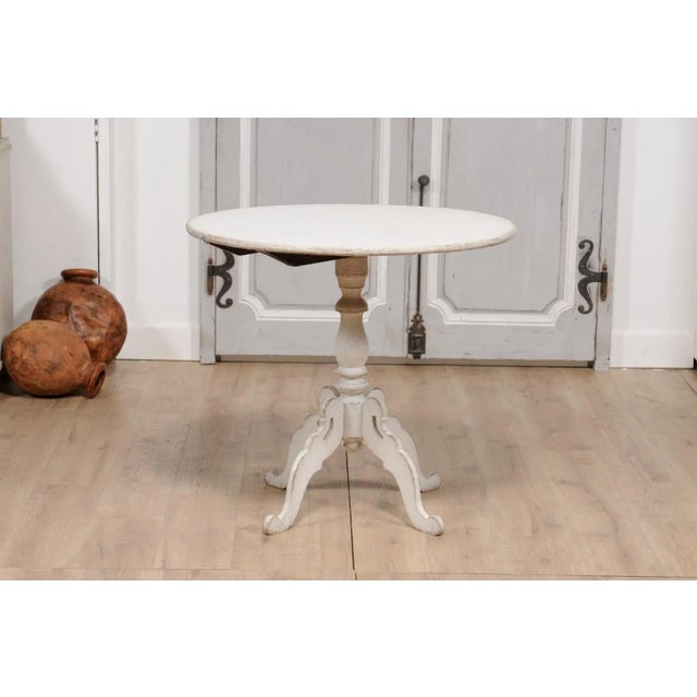 A Swedish light grey painted tilt-top table from circa 1860 with circular top and turned pedestal mounted on four carved...
