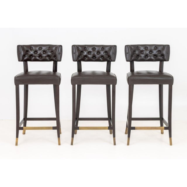 Set of Three Brabbu Chocolate Leather Bar Stools, with four ebonized wood legs conjoined by brass stretchers, label to...